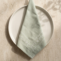 Chikankari sage green linen napkin with white hand embroidered, displayed on a round off-white ceramic plate against a textured beige stone surface with soft leaf shadows.
