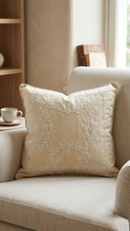 A square beige Chikankari cushion cover pillow with white floral embroidery in the center and a braided rope trim along the edges, shown on a light sofa