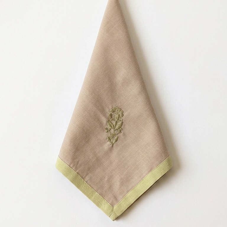 A beige linen napkin with a light green border and a small green chikankari embroidered floral motif displayed against a plain white background.