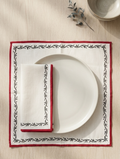A place setting featuring a square chikankari white linen placemat with a red border and black floral vine pattern, styled with a white plate and matching folded napkin on a light wood tabletop.