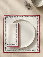 A place setting featuring a square chikankari white linen placemat with a red border and black floral vine pattern, styled with a white plate and matching folded napkin on a light wood tabletop.