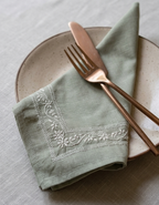 Chikankari sage green linen napkin with white floral hand embroidery, styled on a speckled beige ceramic plate with rose gold-toned flatware.